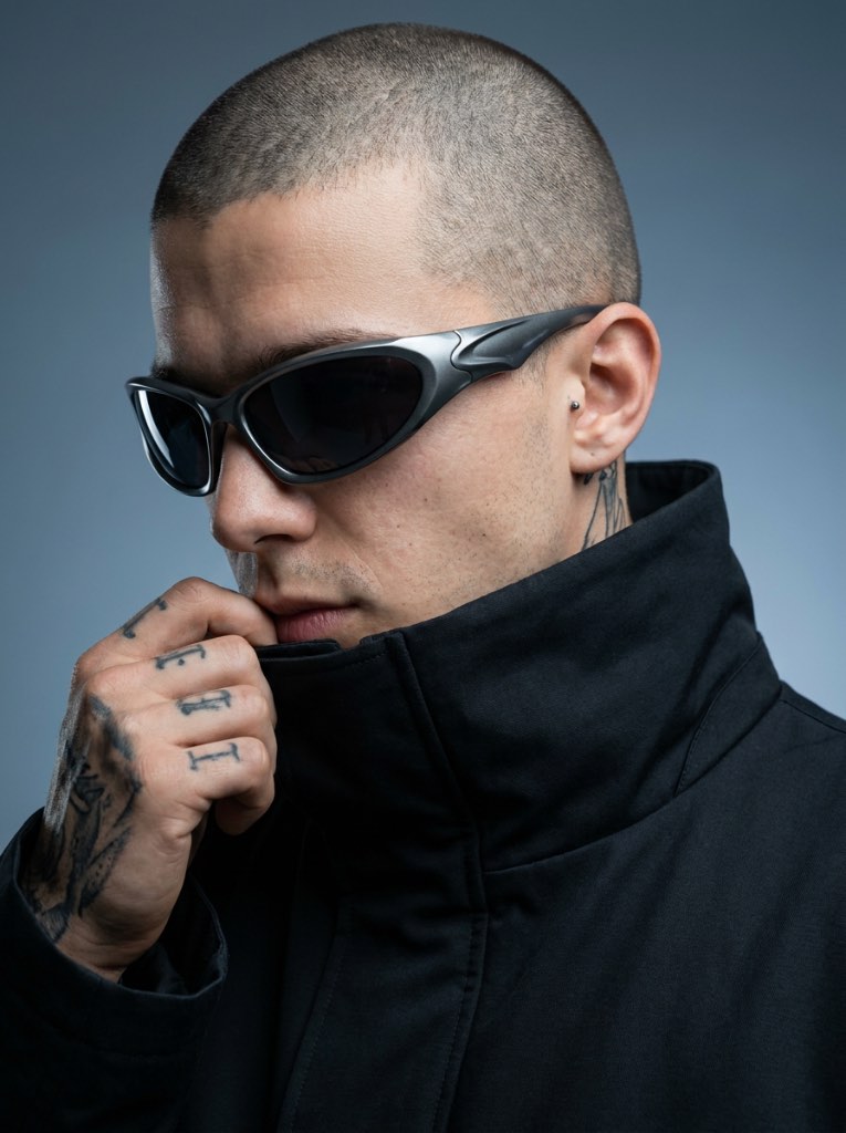 Close-cropped man in wraparound sunglasses holding a high black collar, with tattoos visible on his hand and neck