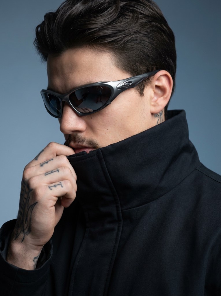 Man with swept-back hair and dark wraparound sunglasses gripping a high black collar with a tattooed hand