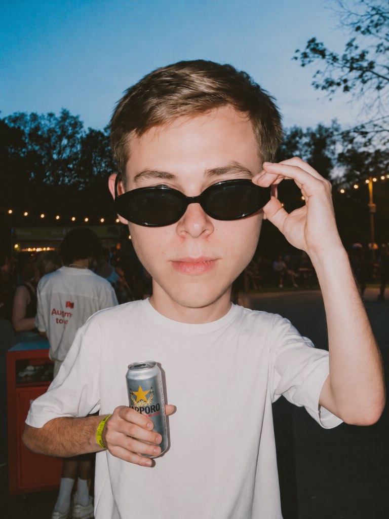 Big-head portrait of a man in a white T-shirt adjusting black sunglasses and holding a drink can outdoors.