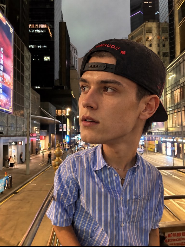 Big-head portrait of a man in a backwards cap and striped shirt above a city street at dusk.