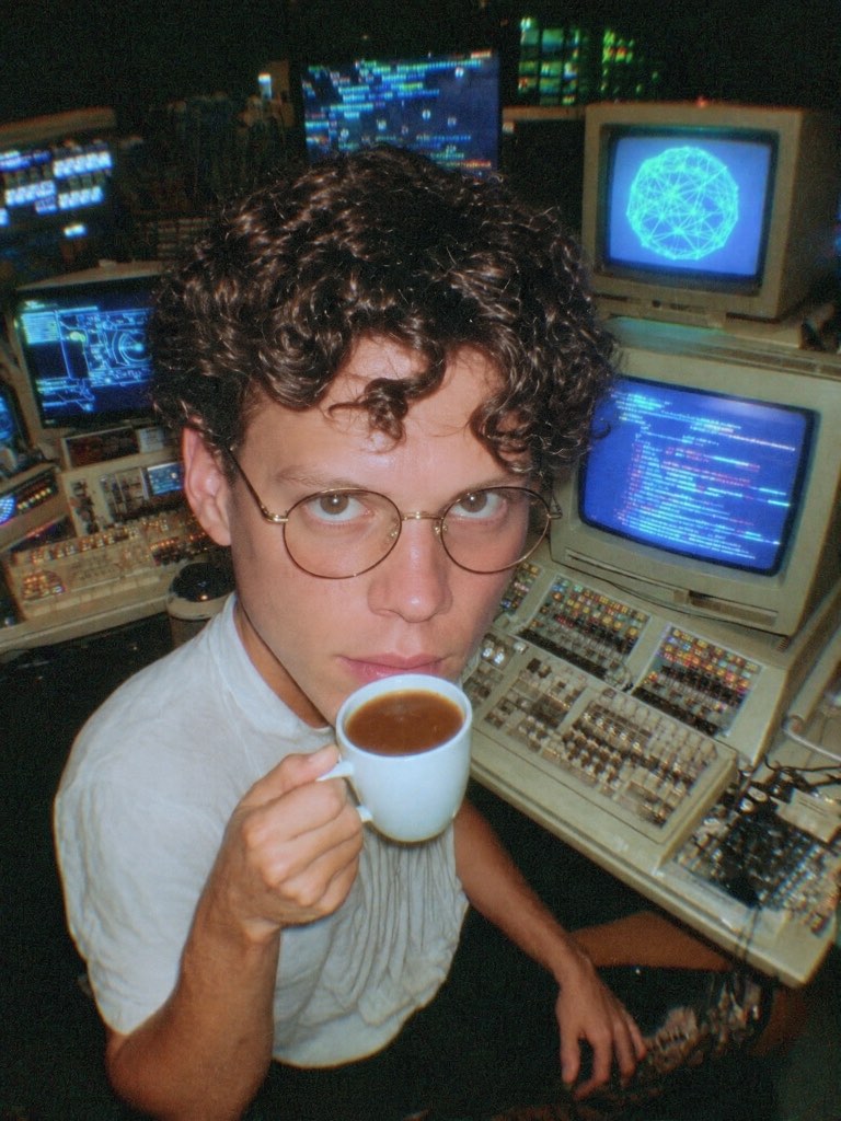 Big-head portrait of a curly-haired man with glasses holding a mug among glowing computer monitors.