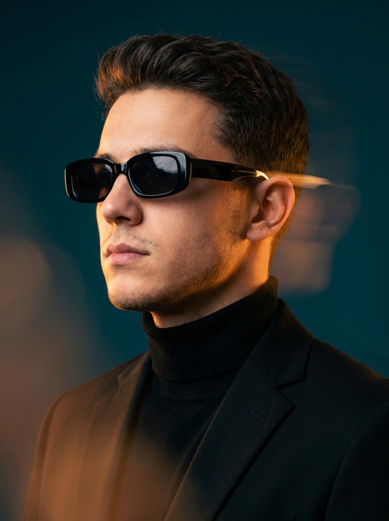 Man with swept-back dark hair and black sunglasses, lit by warm amber light against a teal studio background.