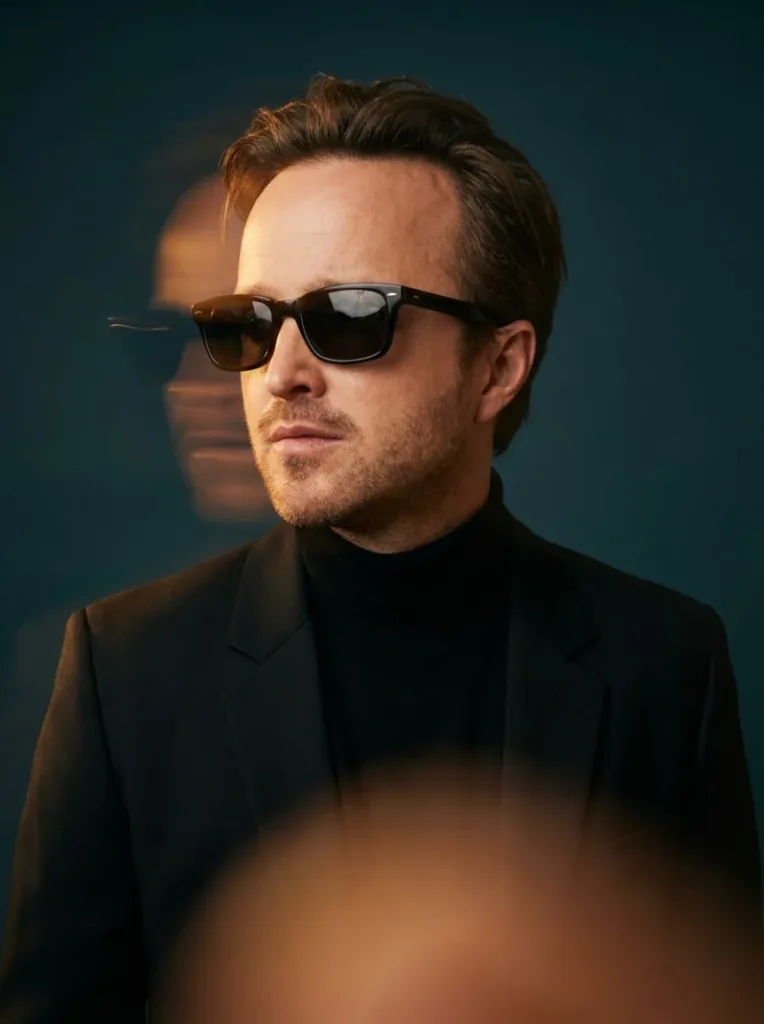 Man in sunglasses and a black turtleneck, with a blurred amber duplicate of his face behind him on a teal background.