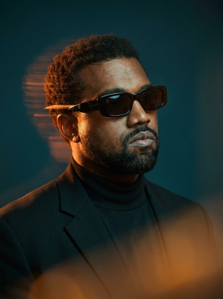 Bearded man in black sunglasses and a blazer, facing right with amber light streaks against a dark teal background.