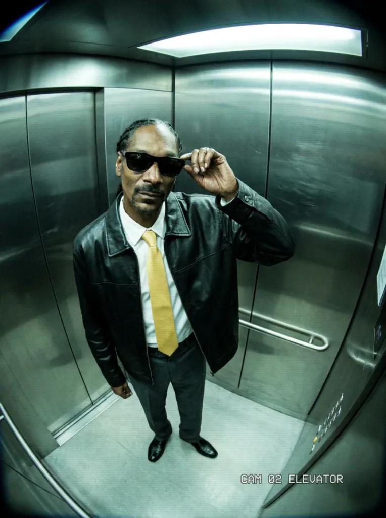 High-angle elevator portrait of a man with braided hair, black sunglasses, a leather jacket and yellow tie.