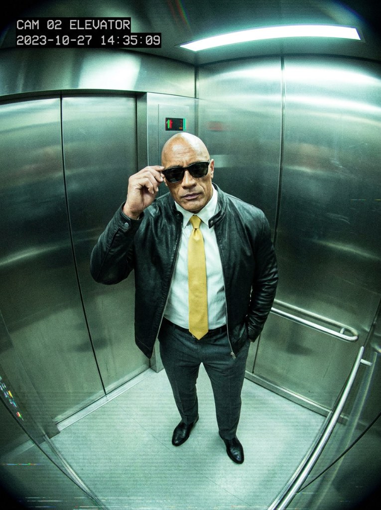 Fisheye elevator view of a bald man leaning toward the camera while adjusting sunglasses above a bright yellow tie.