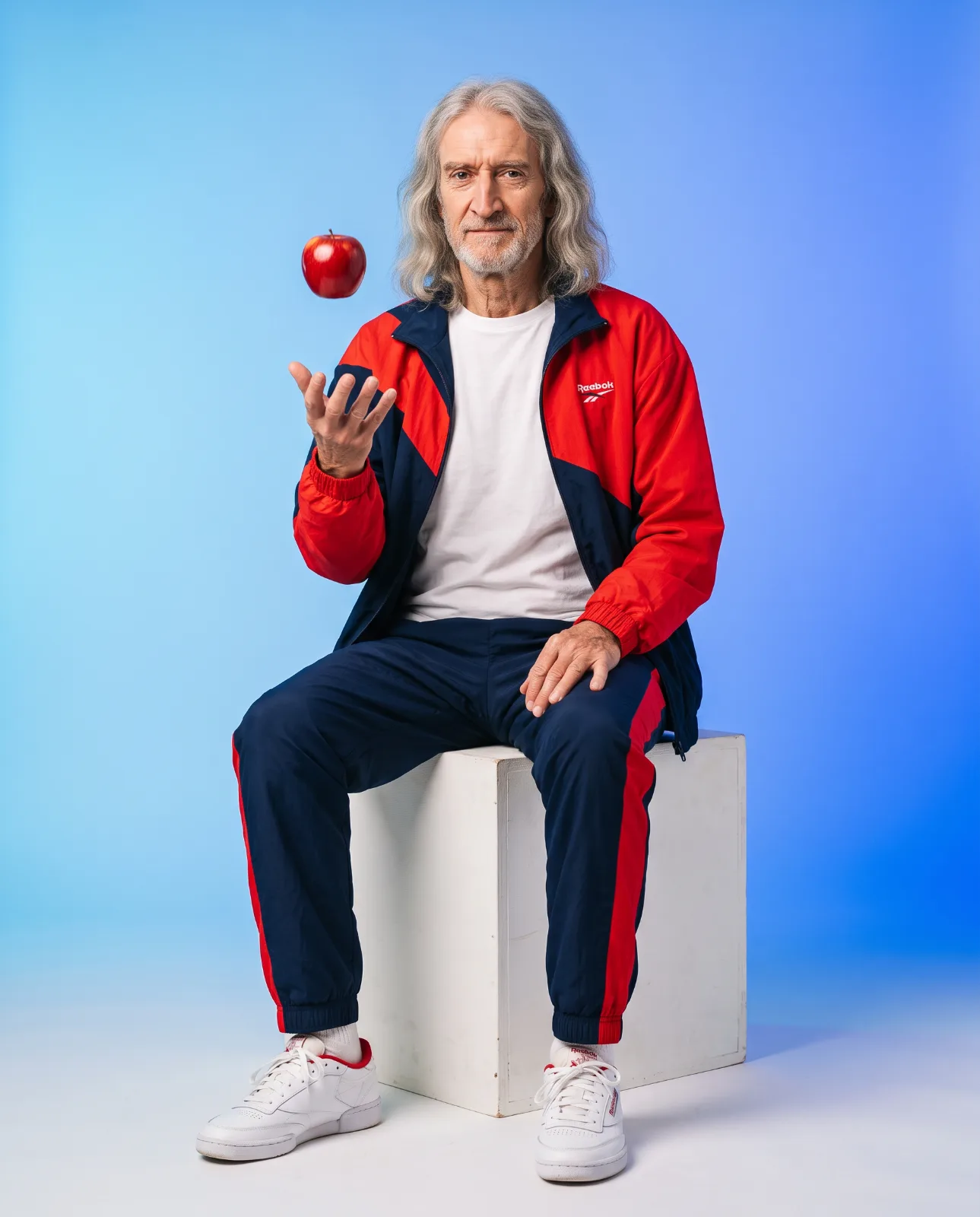Gray-haired man in a red and navy tracksuit seated on a cube with a red apple suspended above his hand