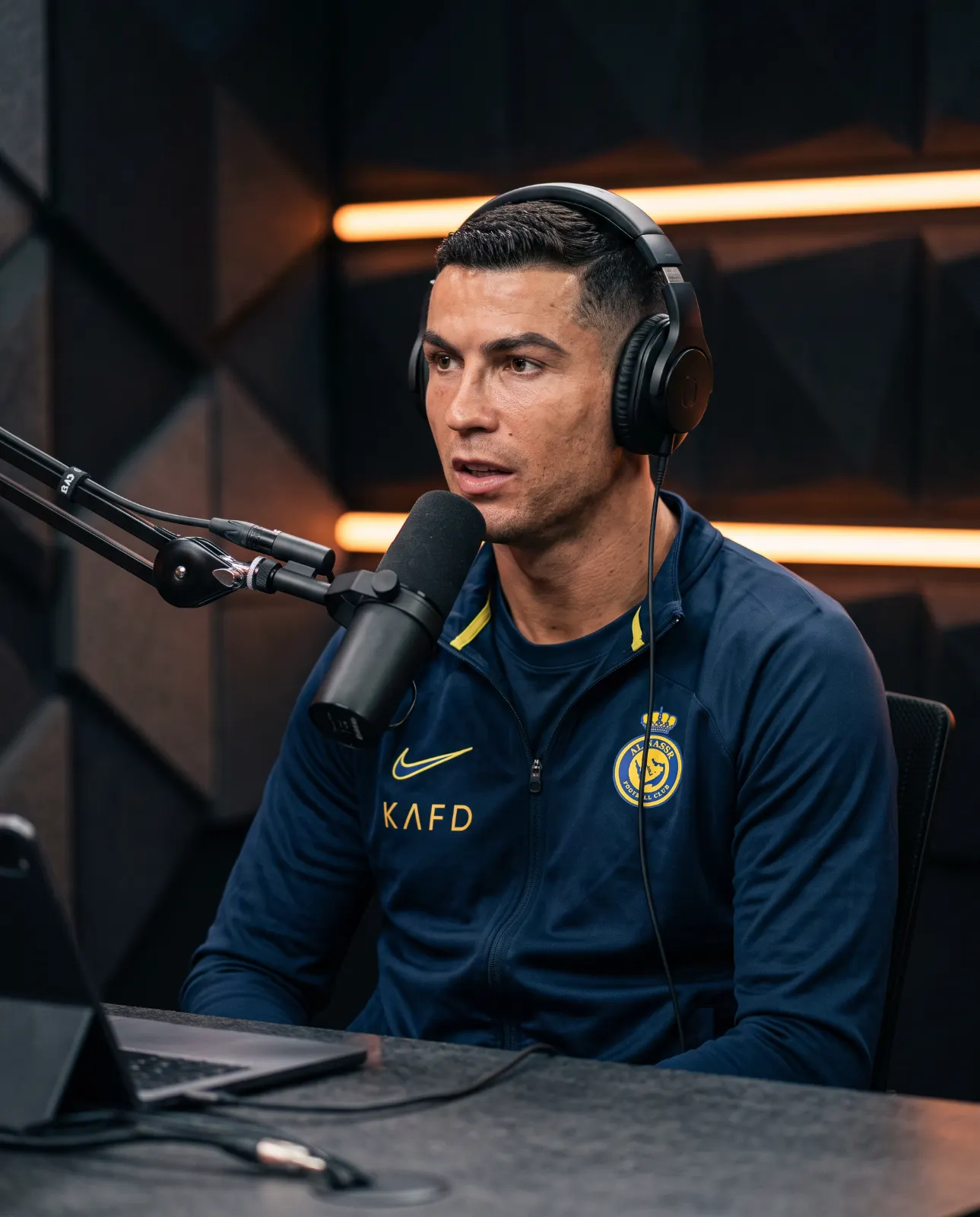 Man in a navy tracksuit and headphones speaking toward a boom microphone beside a laptop