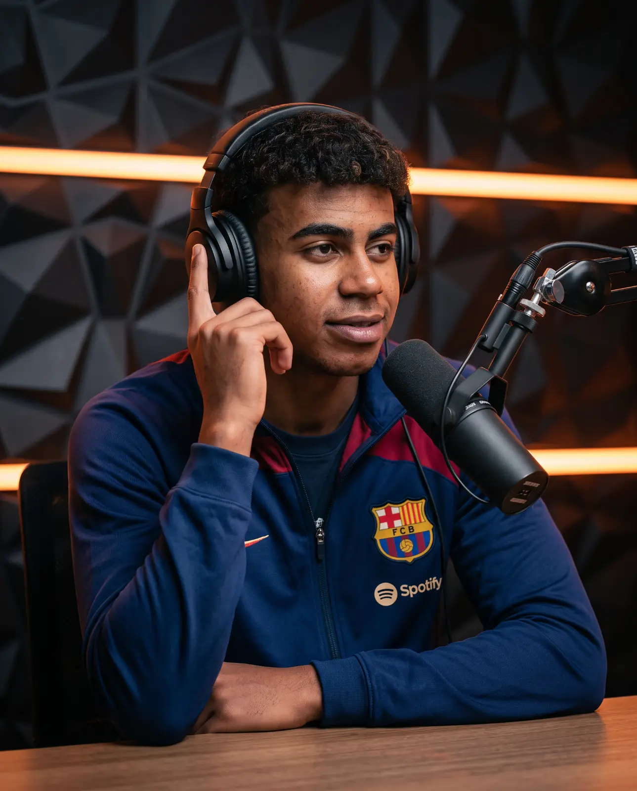 Curly-haired man in a navy football jacket touching his headphones beside a podcast microphone