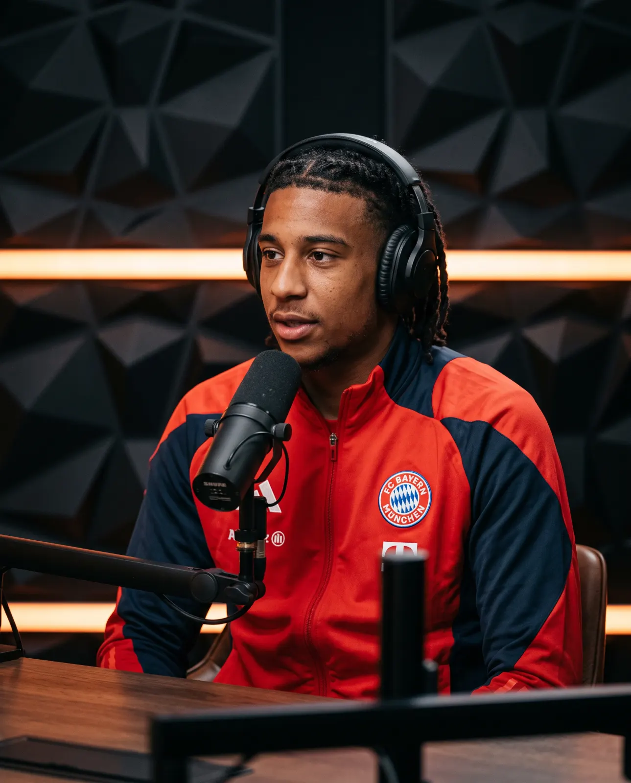 Man with braided hair in a red and navy tracksuit seated at a microphone in a dark podcast studio