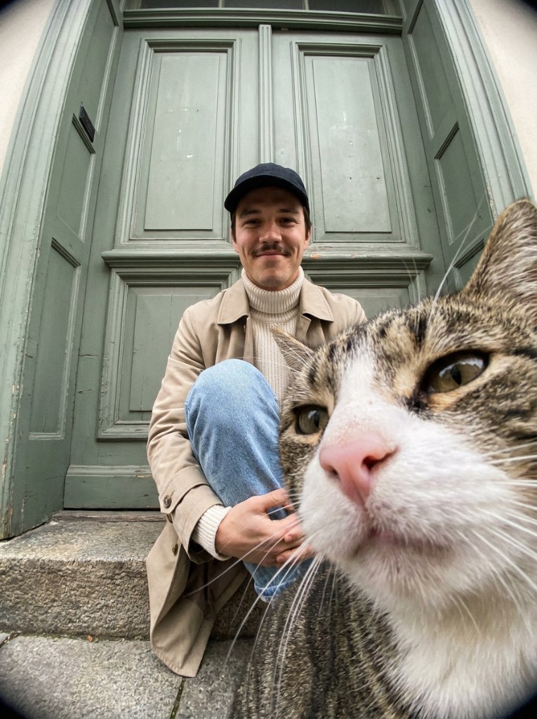 Close fisheye view of a tabby-and-white cat with a smiling man in a black cap seated on a doorstep behind it.