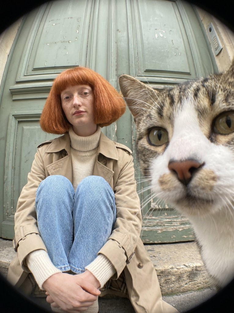 Tabby cat leaning close to a fisheye lens beside a woman with a red bob, seated in front of green wooden doors.