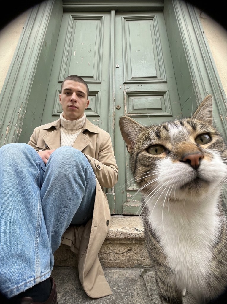 Fisheye doorstep portrait of a short-haired man in a beige coat, with a tabby-and-white cat in the right foreground.